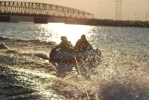 Two people on a jet ski create splashes as they speed across a shimmering water surface at sunset.