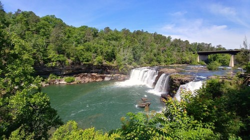 A scenic view of a waterfall cascading into a river, surrounded by lush greenery and a bridge in the background.