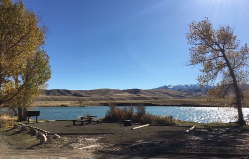 A serene lakeside scene with a picnic area, surrounded by autumn trees and mountains under a clear blue sky.