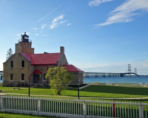 A lighthouse with a red roof beside a calm lake, with a bridge visible in the background under a clear blue sky.