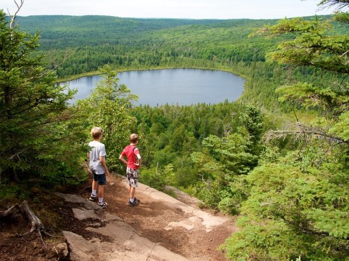 Two boys stand on a rocky ledge overlooking a forested landscape and a serene lake in the distance.
