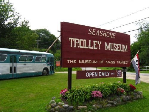 Sign for Seashore Trolley Museum with a vintage bus parked nearby, surrounded by greenery. Open daily.