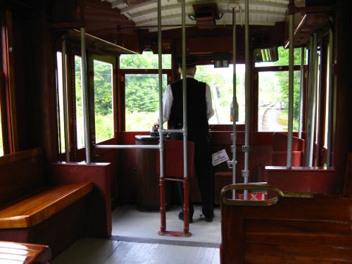 Interior view of a vintage train car, featuring a conductor at the controls and wooden benches. Green scenery outside.