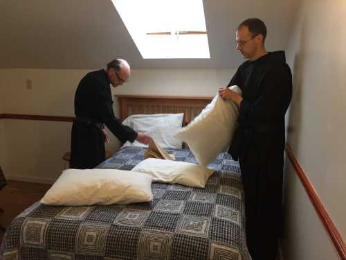 Two men in dark robes are making a bed in a room with a skylight, arranging pillows on a patterned bedspread.