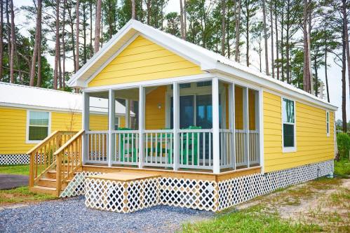 A bright yellow cabin with a porch, surrounded by trees and gravel, showcasing a welcoming outdoor space.