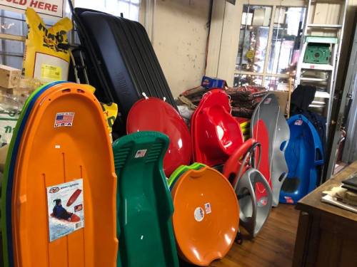 A variety of colorful sleds displayed in a store, with bags and other items in the background.