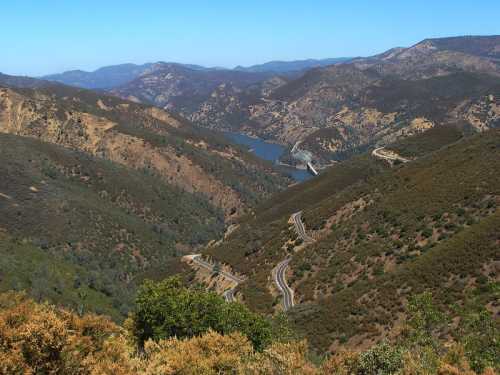 A scenic view of winding roads through green hills, overlooking a river and distant mountains under a clear blue sky.