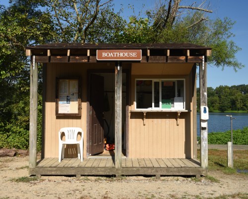 A small wooden boathouse with a sign, featuring a porch and a view of a lake in the background.