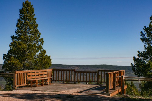 A wooden viewing platform surrounded by trees, overlooking a scenic mountain landscape under a clear blue sky.