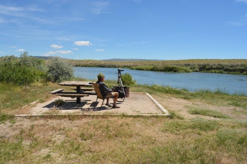 A person sits at a picnic table by a river, surrounded by greenery and under a clear blue sky.