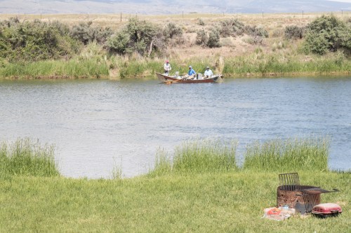 A small boat with three people floats on a calm river, with grassy banks and a grill nearby.