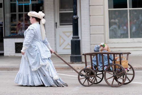 A woman in a vintage blue dress pulls a wooden cart with a child drinking from a can inside.