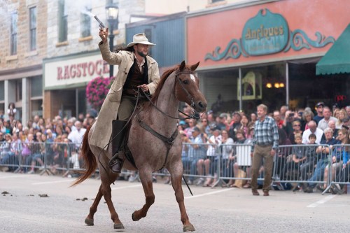A cowboy in a long coat rides a horse, firing a gun, with a crowd watching in a historic town setting.