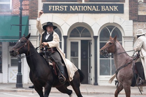 Two cowboys on horseback in front of a bank, one raising a gun, set in a Western-style town.