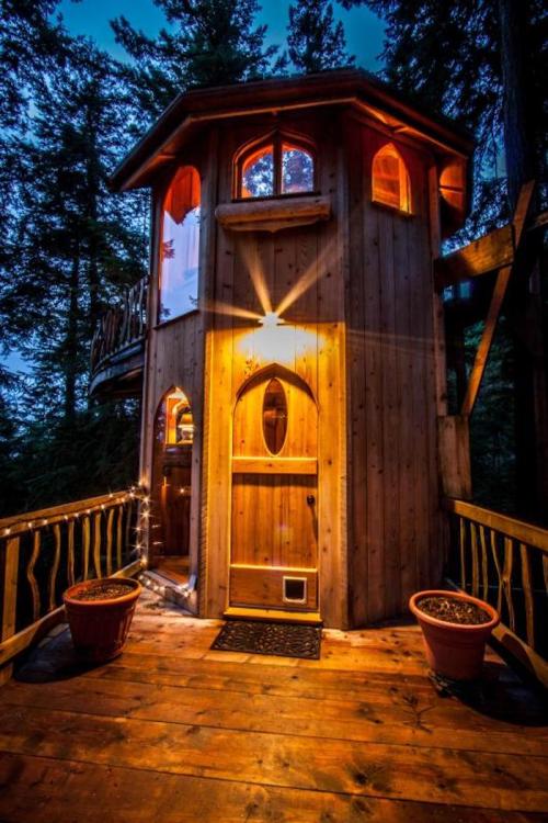 A cozy wooden treehouse illuminated at dusk, surrounded by trees, with a welcoming door and decorative lights.
