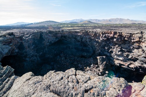 A vast volcanic landscape with a large crater and rocky terrain, surrounded by distant mountains under a clear sky.