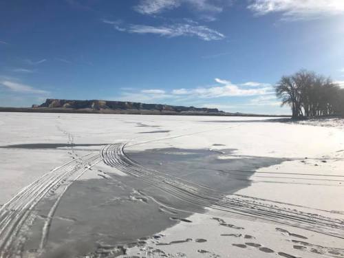 A snowy landscape with tire tracks on a frozen lake, surrounded by cliffs and trees under a clear blue sky.