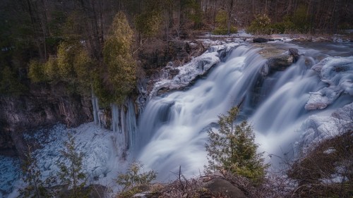 A serene waterfall cascades over rocks, surrounded by trees and a misty atmosphere in a tranquil natural setting.