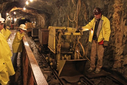 A miner demonstrates equipment in a dimly lit underground tunnel, with visitors observing in safety gear.