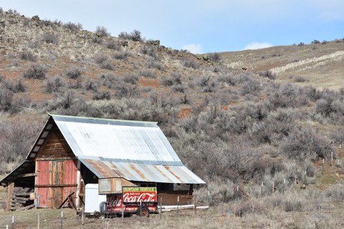 A rustic barn with a metal roof and a vintage Coca-Cola trailer, set against a hilly, shrub-covered landscape.