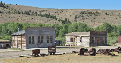 Two old wooden buildings in a rural landscape, surrounded by hills and remnants of vintage machinery.
