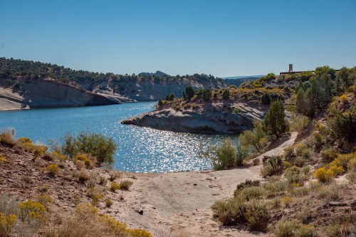 A serene lake surrounded by rocky shores and greenery under a clear blue sky. Sunlight sparkles on the water's surface.
