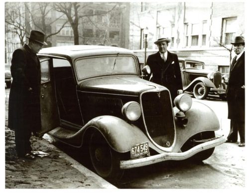 Two men in hats stand beside a vintage car parked on a city street, with another car visible in the background.
