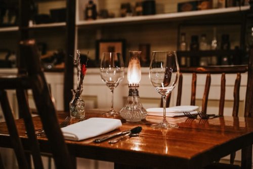 A cozy dining table set with wine glasses, a lit oil lamp, and elegant tableware in a warm, inviting atmosphere.