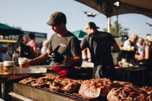 Two chefs grill large cuts of meat at a busy outdoor food festival, with a crowd and tents in the background.