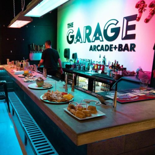 A vibrant bar with colorful lighting, featuring a bartender, drinks, and a variety of appetizers on the counter.