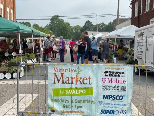 A bustling outdoor market with vendors and shoppers, featuring tents and a banner for the Valparaiso Market.