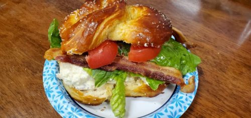 A croissant sandwich filled with lettuce, tomato, bacon, and creamy filling, served on a paper plate.