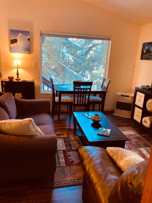Cozy living area with a sofa, coffee table, and dining space by a window overlooking trees. Warm, inviting atmosphere.