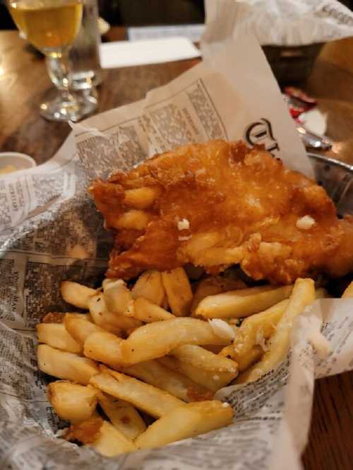 A basket of crispy fried fish and golden French fries, served on newspaper. A drink is visible in the background.