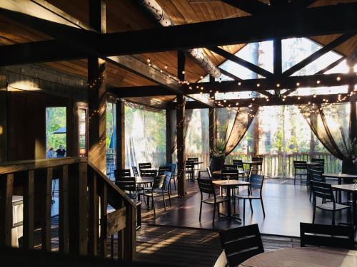 A rustic dining area with wooden beams, string lights, and empty tables, surrounded by large windows and greenery.