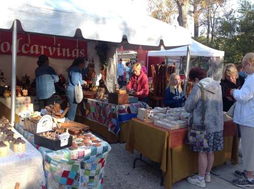 A craft fair scene with people browsing wooden carvings and handmade items under a large tent.