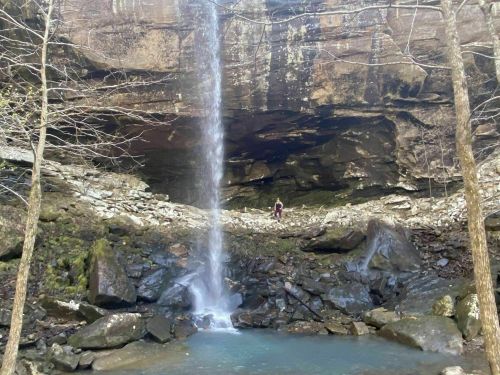 A waterfall cascades over rocky terrain, with a person standing beneath the cliff in a serene natural setting.