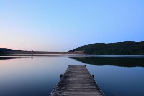A calm lake at dusk with a wooden dock extending into the water, surrounded by hills and a dam in the distance.
