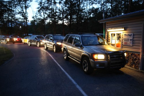 Cars lined up in a queue outside a small food stand at dusk, surrounded by trees.