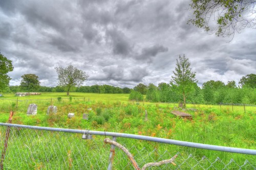 A cloudy sky over a grassy field with a fenced area containing scattered gravestones and trees.
