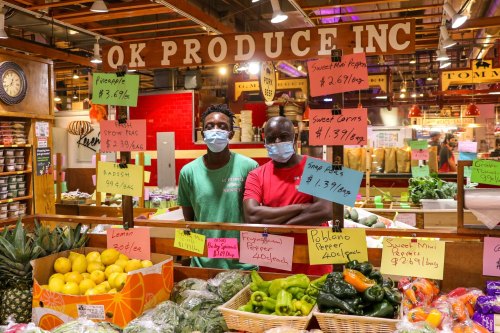 Two men wearing masks stand behind a produce stand filled with colorful fruits and vegetables, with price signs visible.