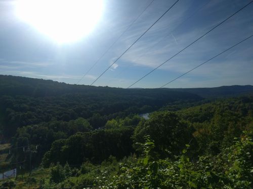 A scenic view of a lush green valley with a river, under a bright sun and blue sky, with power lines in the foreground.