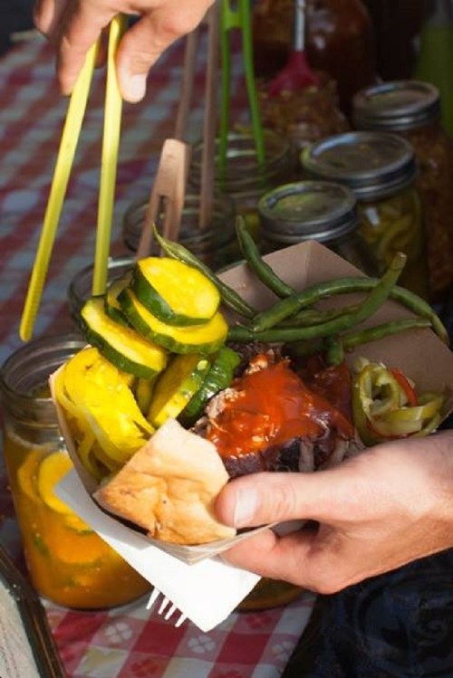 A hand holds a bowl of food with pickles, green beans, and sauce, with jars of pickled vegetables in the background.