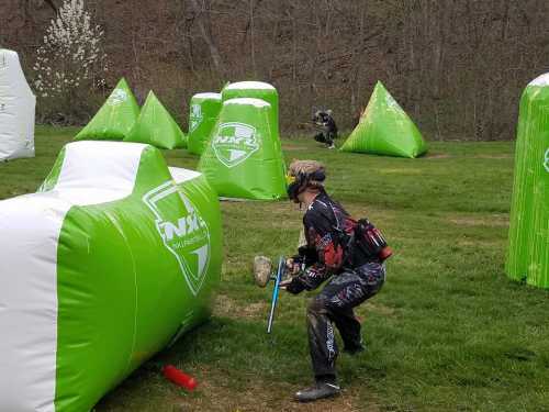 A player crouches behind a green inflatable barrier on a paintball field, aiming with a paintball marker.