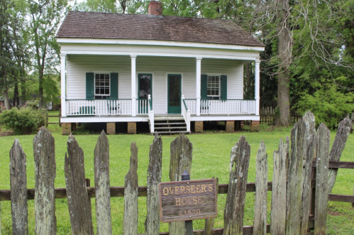 A historic white house with green shutters, surrounded by a wooden fence, labeled "Overseer's House, 1870."