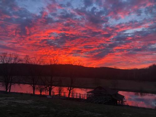 A vibrant sunset with red and orange clouds reflecting on a calm lake, framed by trees and distant hills.