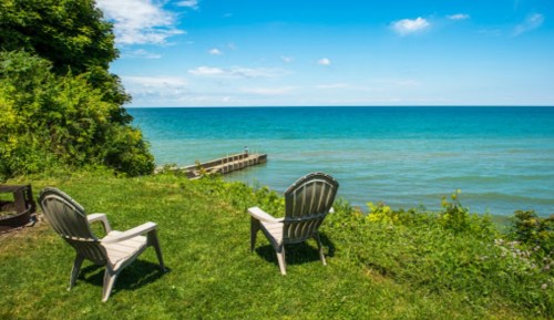 Two chairs facing a calm blue lake, surrounded by green grass and trees under a clear sky.