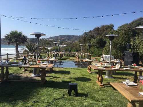 Outdoor dining setup with wooden tables, heaters, and ocean view, surrounded by greenery and string lights.