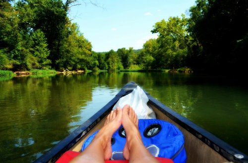 A person's feet resting on the edge of a canoe, surrounded by calm water and lush green trees.