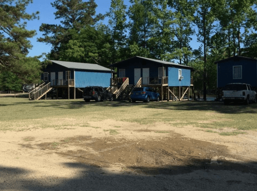 Two blue cabins on stilts surrounded by trees, with parked cars in front and a grassy area nearby.